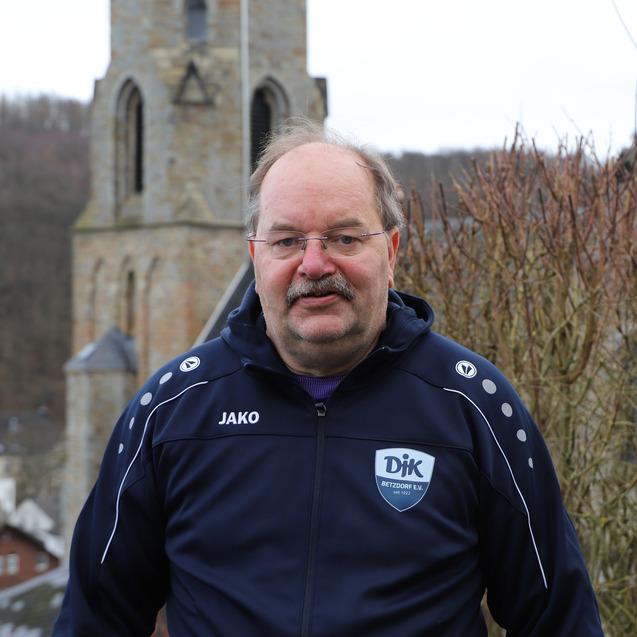 Mann in Sportjacke steht vor einer Kirche mit Turm und Blick auf eine l&auml;ndliche Umgebung.