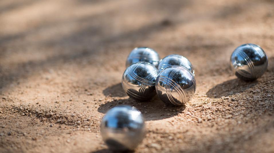 Sechs glänzende Metallkugeln liegen im Sand eines Boule-Platzes.