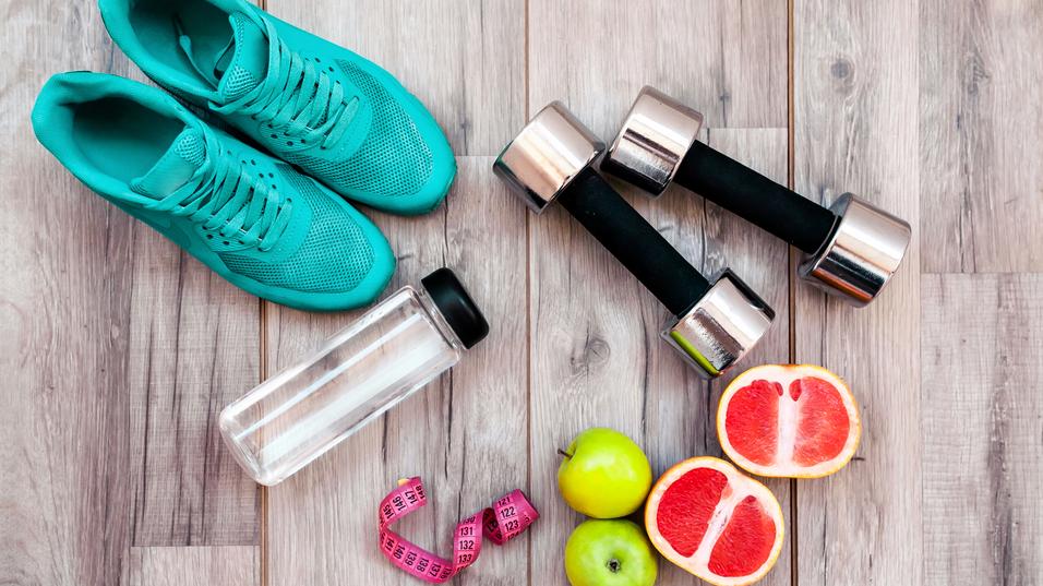 Grüne Sportschuhe, zwei Hanteln, eine Wasserflasche, eine Maßband und frisches Obst auf einem Holzboden.