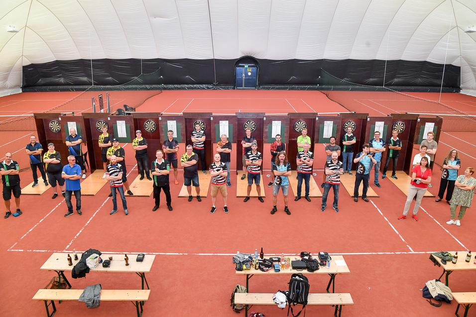 Gruppe von Dartspielern steht auf einer roten Indoor-Tennishalle zwischen Dartboards und Tischen.