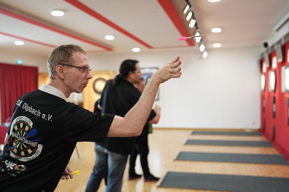 Mann in Sportshirt wirft Dartpfeil auf eine Dartscheibe in einem gut beleuchteten Spielraum.