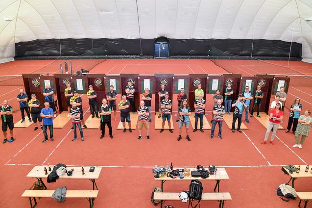Gruppe von Dartspielern steht auf einer roten Indoor-Tennishalle zwischen Dartboards und Tischen.