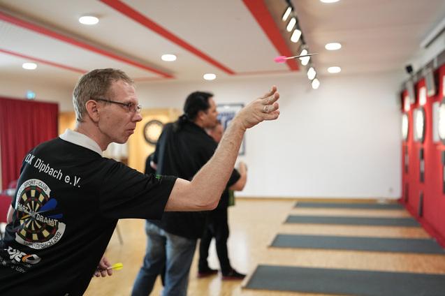 Mann in Sportshirt wirft Dartpfeil auf eine Dartscheibe in einem gut beleuchteten Spielraum.