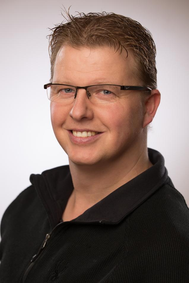 Portr&auml;t eines l&auml;chelnden Mannes mit Brille, der einen schwarzen Pullover tr&auml;gt, vor einem hellen Hintergrund.