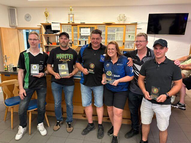 Sechs Personen halten Pokale in einem Clubraum mit Regalen im Hintergrund. Sie posieren l&auml;chelnd vor einer Theke.