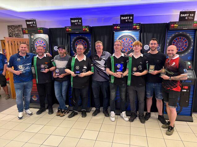 Gruppierung von acht Dartspielern mit Pokalen vor Dartautomaten in einem Spielraum.