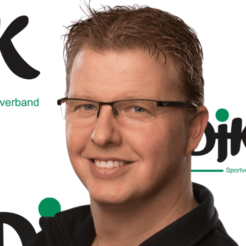 Portr&auml;t eines l&auml;chelnden Mannes mit Brille, in einem schwarzen Oberteil, vor einem unscharfen Hintergrund mit Logos.