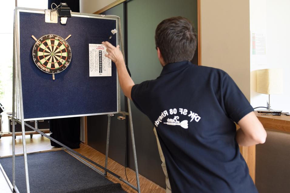 Mann wirft einen Dartpfeil auf eine Dartscheibe in einem Innenraum mit Holzfu&szlig;boden.