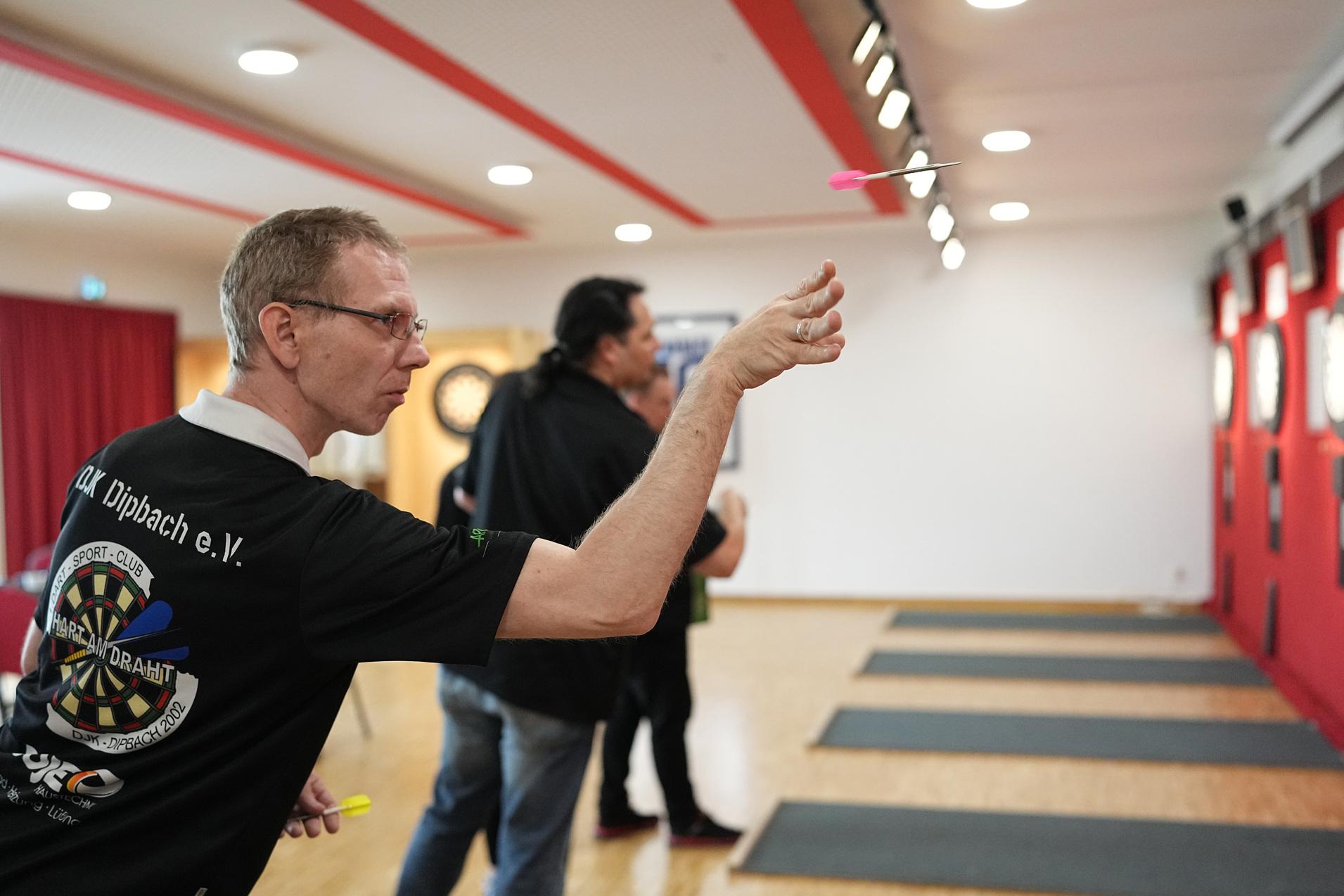 Mann in Sportshirt wirft Dartpfeil auf eine Dartscheibe in einem gut beleuchteten Spielraum.