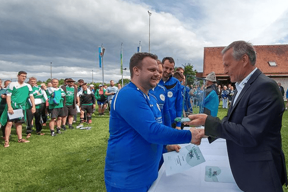 Ein Spieler in blauer Sportkleidung erh&auml;lt einen Pokal w&auml;hrend einer Ehrungsveranstaltung auf einem Rasenplatz.