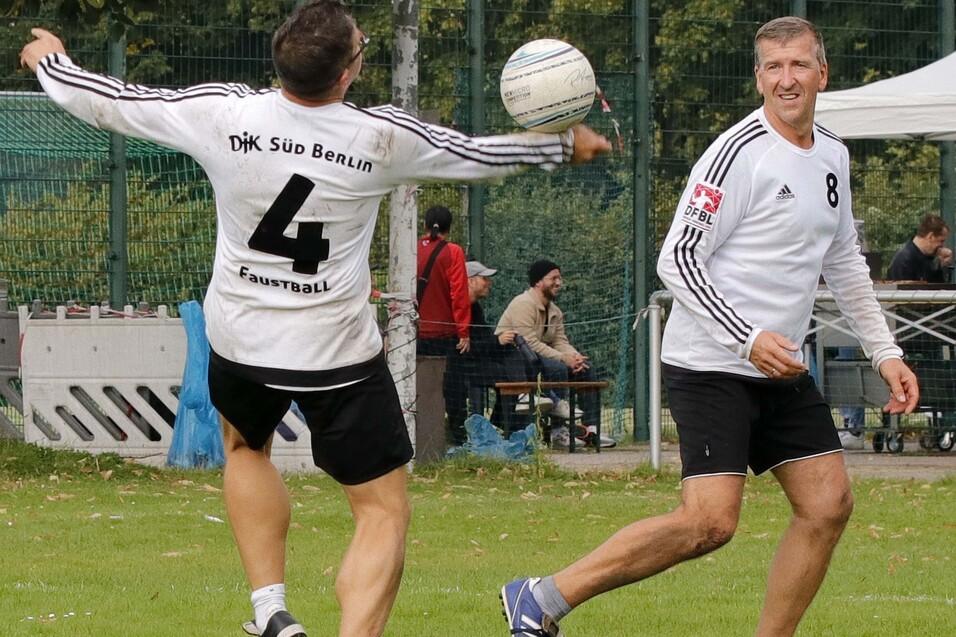 Zwei Faustballspieler im wei&szlig;en Trikot, auf einem Sportfeld in Berlin, werfen einen Ball zueinander.