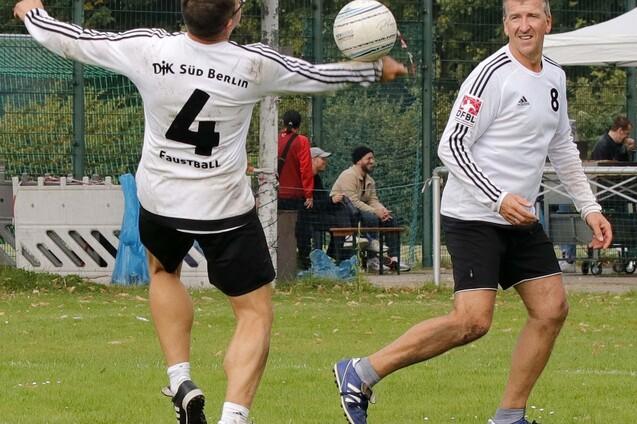 Zwei Faustballspieler im wei&szlig;en Trikot, auf einem Sportfeld in Berlin, werfen einen Ball zueinander.