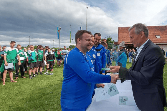 Ein Spieler in blauer Sportkleidung erh&auml;lt einen Pokal w&auml;hrend einer Ehrungsveranstaltung auf einem Rasenplatz.