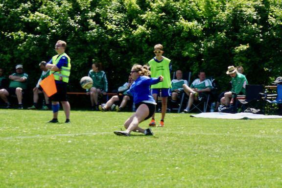 Eine Spielerin in blauer Sportkleidung schie&szlig;t einen Fu&szlig;ball, w&auml;hrend Zuschauer im Hintergrund auf einer Wiese sitzen.