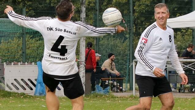 Zwei Faustballspieler im wei&szlig;en Trikot, auf einem Sportfeld in Berlin, werfen einen Ball zueinander.