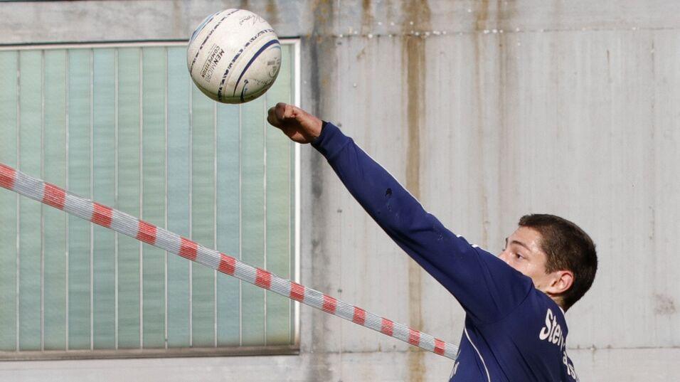Ein Spieler springt und wirft einen Ball über das Spielfeld, während Zuschauer im Hintergrund sitzen.