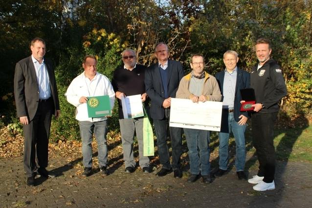 Gruppenfoto von sieben M&auml;nnern im Freien, die Auszeichnungen und einen gro&szlig;en Scheck halten. Herbstliche Natur im Hintergrund.