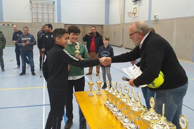 Junge erh&auml;lt einen Pokal von einem Erwachsenen in einer Sporthalle, Zuschauer beobachten die Zeremonie.