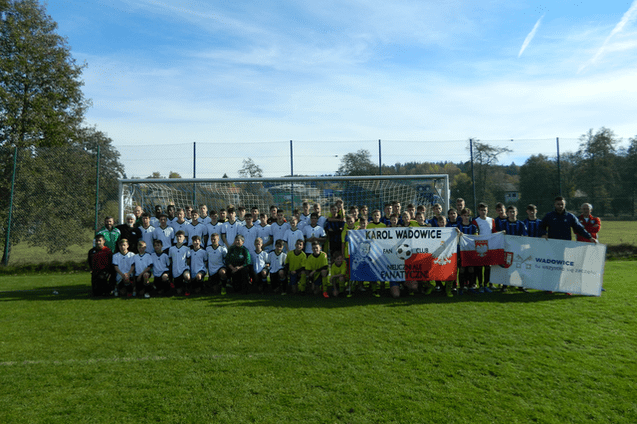 Gruppenfoto von Fu&szlig;ballspielern in wei&szlig;en und schwarzen Trikots vor einem Tor auf einem Sportplatz, Banner im Vordergrund.