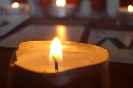 Nahaufnahme einer brennenden Kerze mit Flamme, im Hintergrund weitere Kerzen und ein Tisch mit Karten.