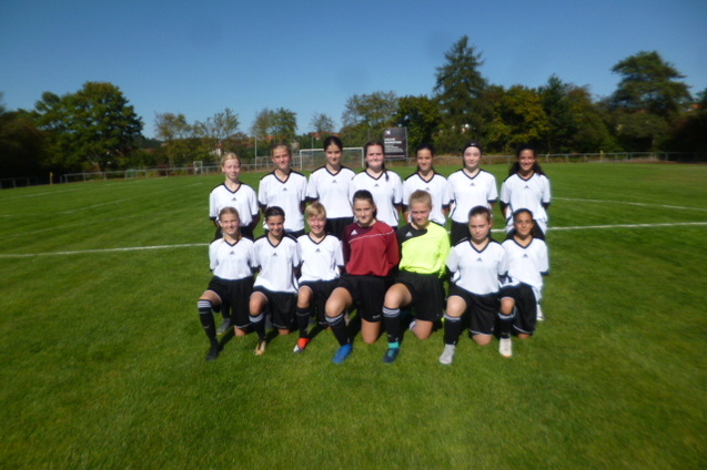 M&auml;dchenfu&szlig;ballmannschaft in einheitlichen Trikots posiert auf einem Sportplatz vor blauem Himmel.