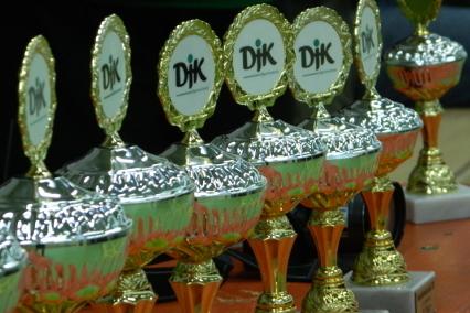 Reihe von goldenen Pokalen mit gr&uuml;n-wei&szlig;en Logos auf einer Tischplatte.