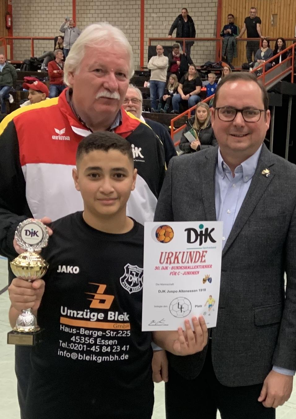 Ein Junge h&auml;lt einen Pokal und eine Urkunde, umgeben von zwei M&auml;nnern in Sportkleidung und Anzug in einer Sporthalle.