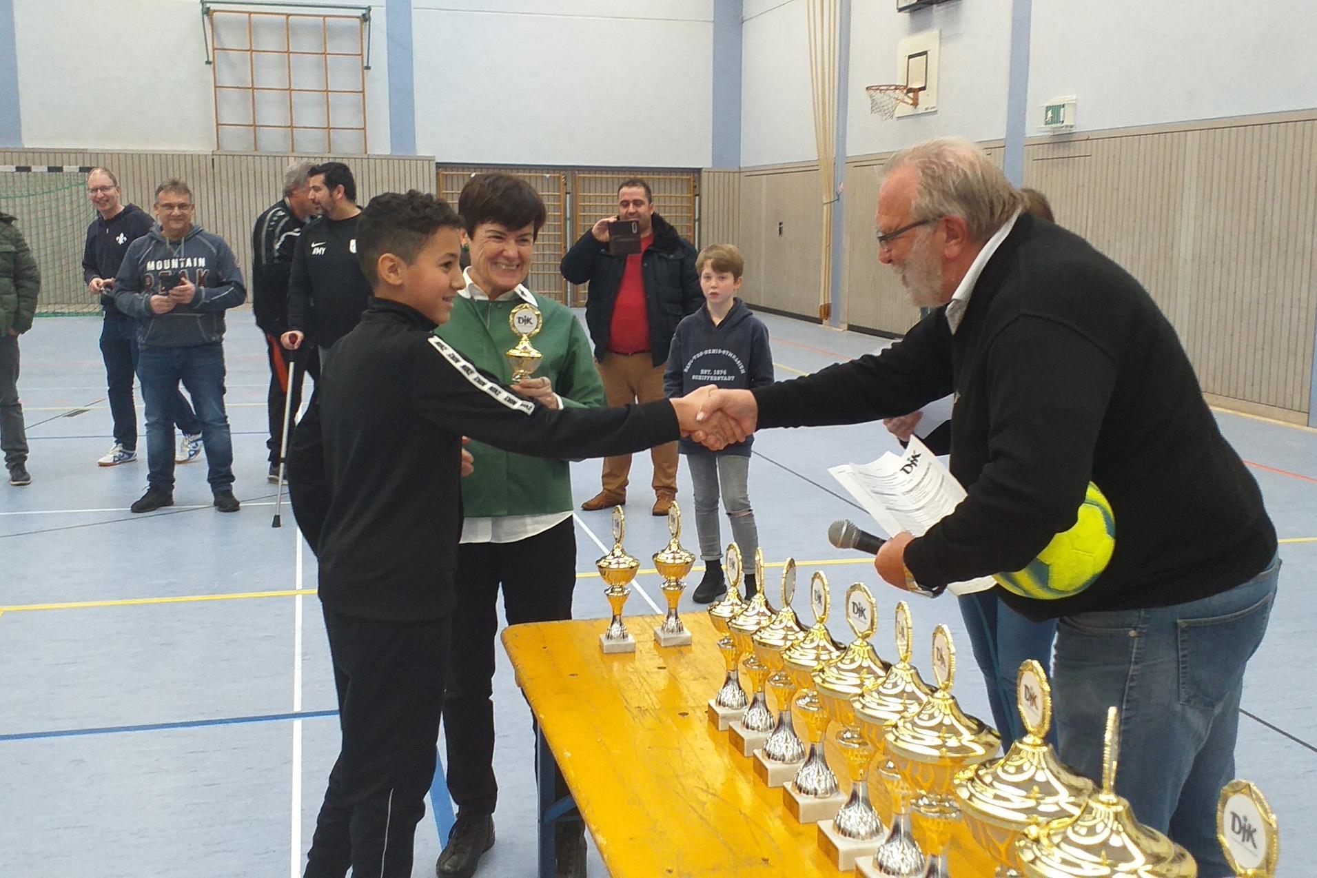 Junge erh&auml;lt einen Pokal von einem Erwachsenen in einer Sporthalle, Zuschauer beobachten die Zeremonie.