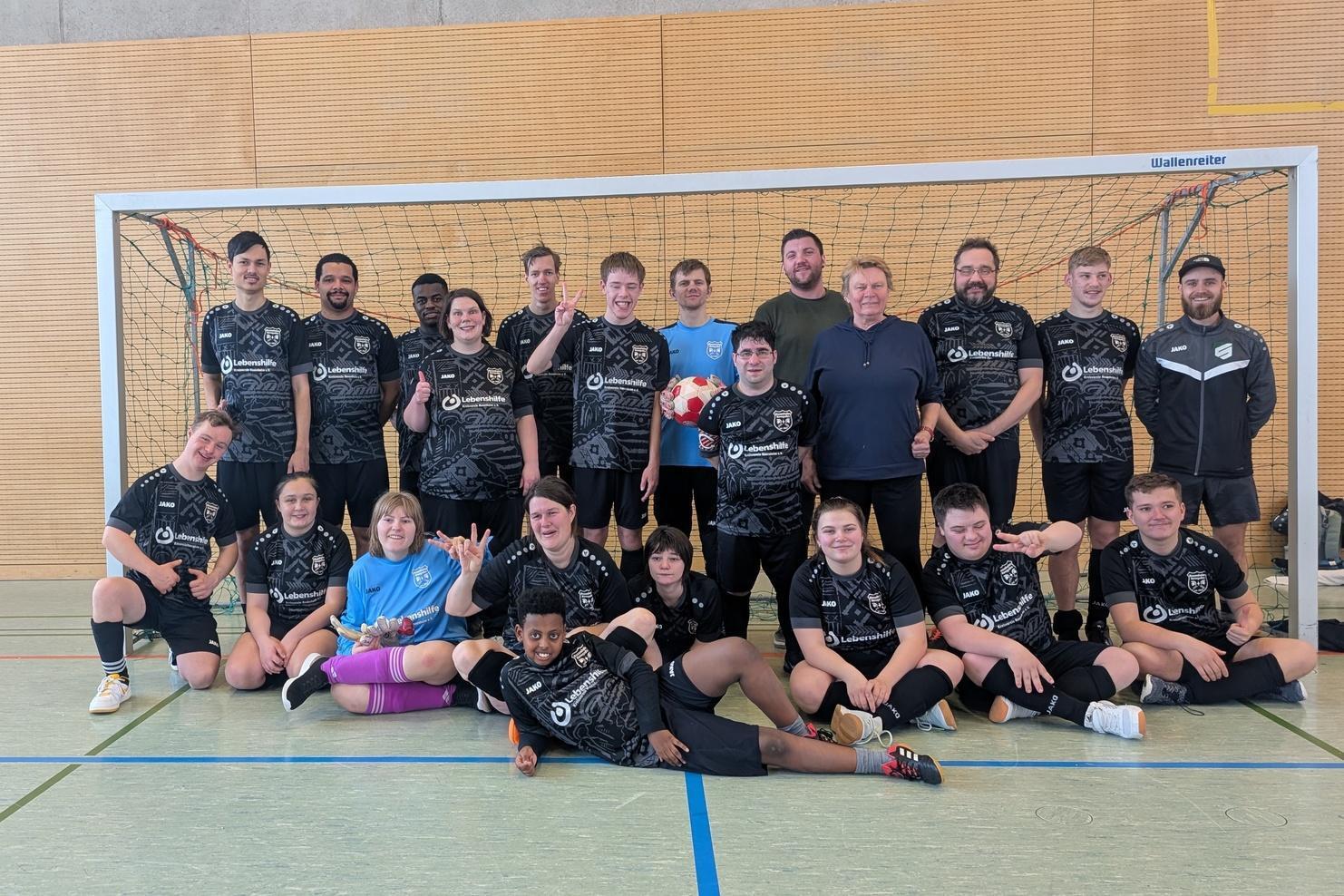 Gruppenshot einer Fu&szlig;ballmannschaft in schwarzen Trikots vor einem Tor in einer Sporthalle.