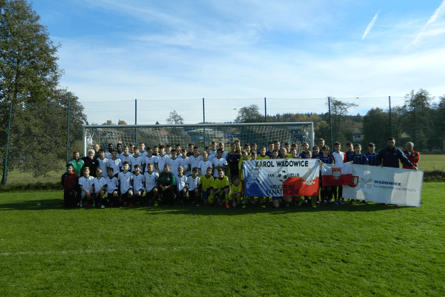 Gruppenfoto von Fu&szlig;ballspielern in wei&szlig;en und schwarzen Trikots vor einem Tor auf einem Sportplatz, Banner im Vordergrund.