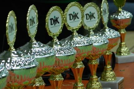 Reihe von goldenen Pokalen mit gr&uuml;n-wei&szlig;en Logos auf einer Tischplatte.