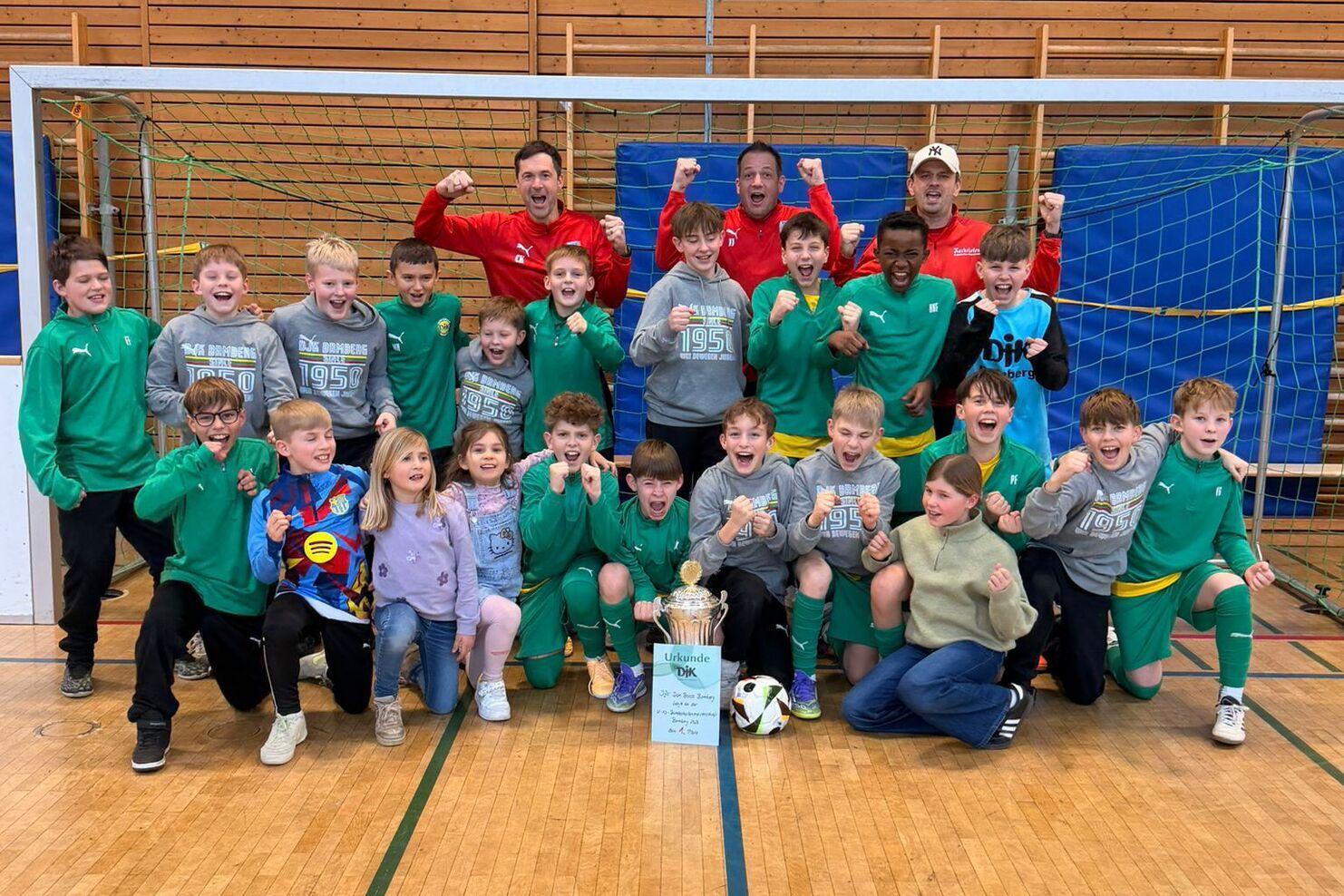Gruppe von Kindern und Trainern feiert vor einem Fu&szlig;balltor mit Pokal und Fu&szlig;ball in einer Sporthalle.