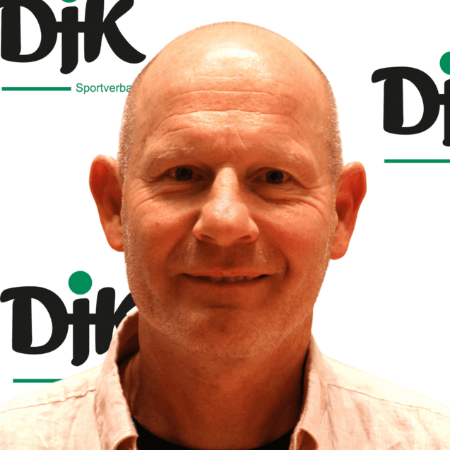 Portr&auml;t eines Mannes mit glattem Kopf und kurzem Hemd, vor einem Hintergrund mit dem Logo des DJK Sportverbands.