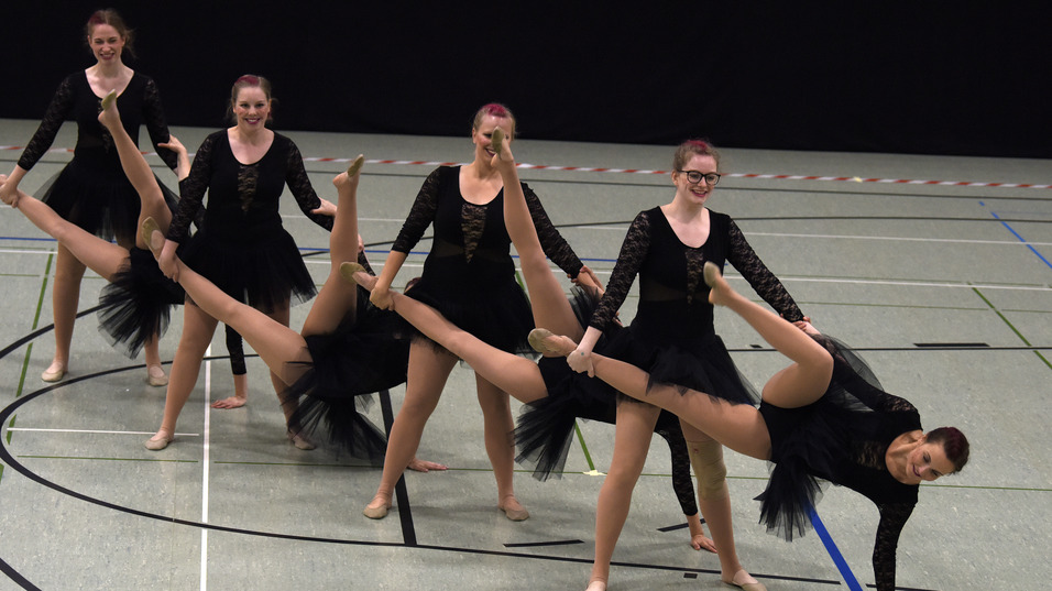Gruppe von T&auml;nzerinnen in schwarzen Kost&uuml;men mit T&uuml;llr&ouml;cken f&uuml;hrt synchronisierte Bewegungen in einer Sporthalle aus.