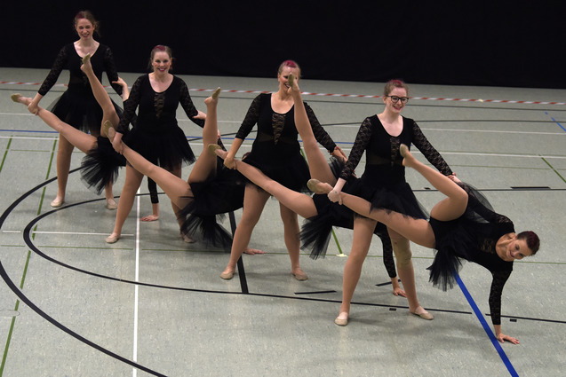 Gruppe von T&auml;nzerinnen in schwarzen Kost&uuml;men mit T&uuml;llr&ouml;cken f&uuml;hrt synchronisierte Bewegungen in einer Sporthalle aus.