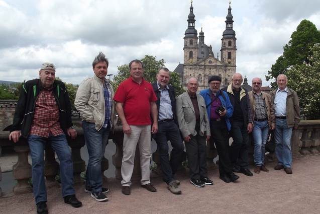 Gruppe von neun M&auml;nnern posiert vor einer zweigeteilten Kirche unter einem bew&ouml;lkten Himmel.