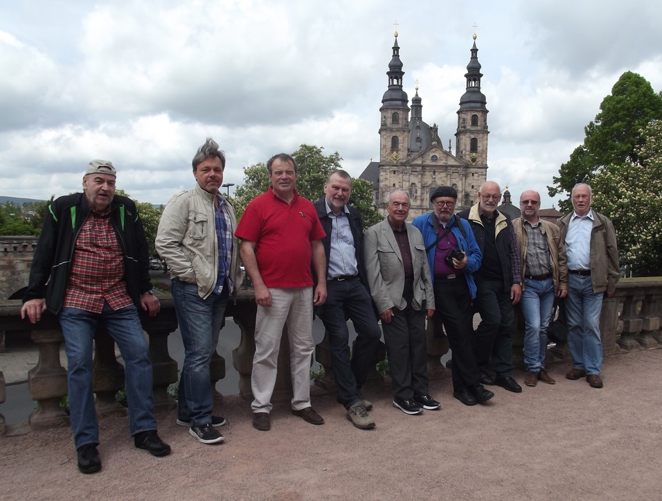 Gruppe von neun M&auml;nnern posiert vor einer zweigeteilten Kirche unter einem bew&ouml;lkten Himmel.
