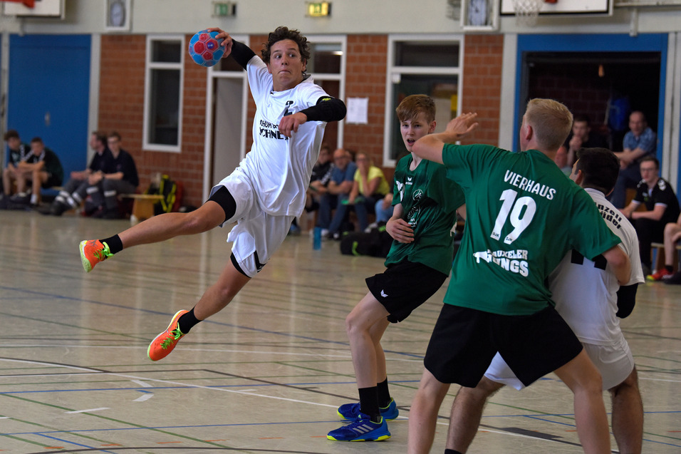 Aktion im Handballspiel: Spieler springt und wirft den Ball, w&auml;hrend Gegner ihn blockieren. Zuschauer im Hintergrund.