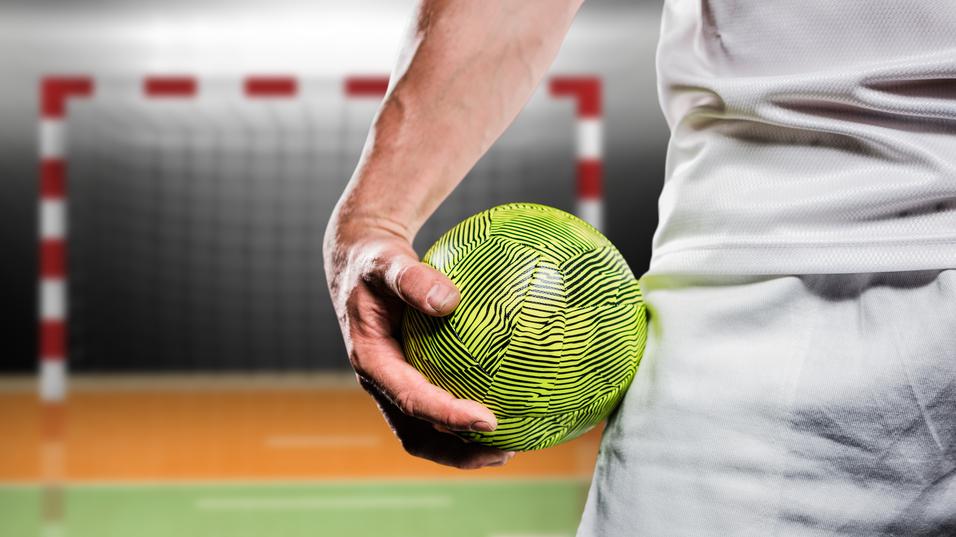 Nahaufnahme einer Hand, die einen grünen Handball hält, im Hintergrund ein Tor auf einem Sportfeld.