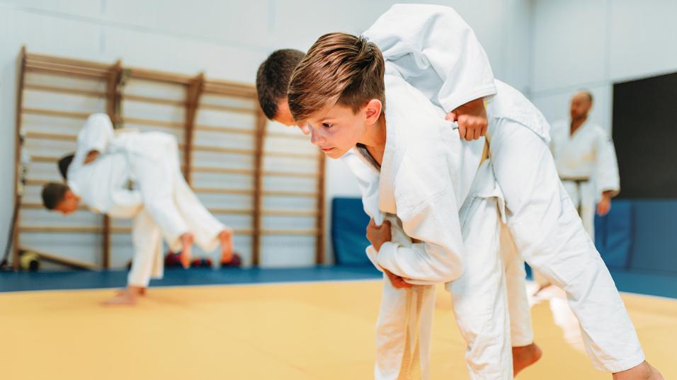 Zwei Kinder in weißen Judo-Anzügen üben einen Wurf auf einer gelben Judomatte. Hintergrund: Weitere Judo-Praktizierende.