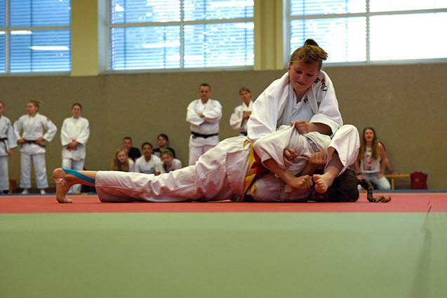 Zwei Judoka im Wettkampf auf einer Matte, eine Athletin kontrolliert die andere in einem Hebel. Zuschauer im Hintergrund.
