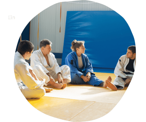 Vier Personen in Judokleidung sitzen im Kreis auf einer Matte, w&auml;hrend sie miteinander sprechen. Ein blauer Mattenbereich im Hintergrund.