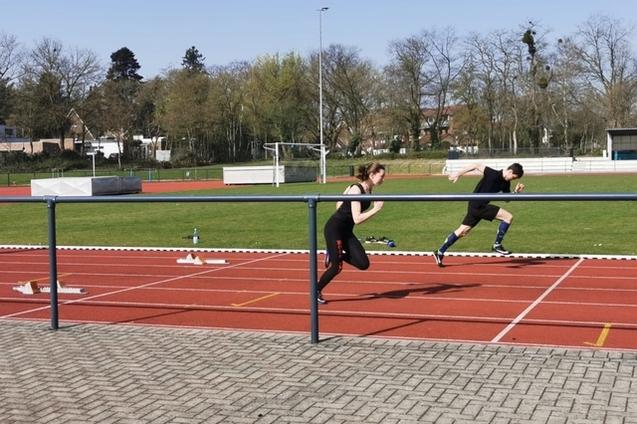 Zwei Athleten sprinten auf einer roten Laufbahn in einem Stadion, umgeben von gr&uuml;nen B&auml;umen und Sporteinrichtungen.