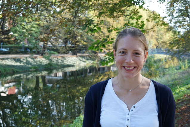 Frau l&auml;chelnd vor einem ruhigen Gew&auml;sser, umgeben von herbstlichen B&auml;umen und Str&auml;uchern.