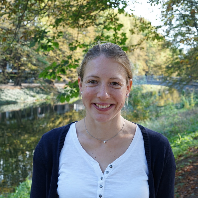 Frau l&auml;chelnd vor einem ruhigen Gew&auml;sser, umgeben von herbstlichen B&auml;umen und Str&auml;uchern.