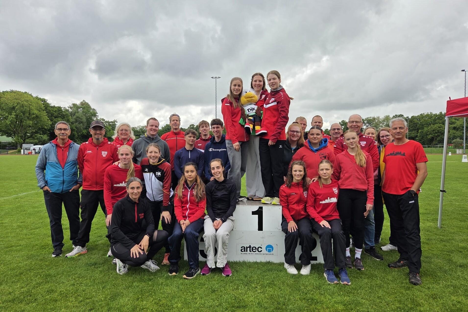 Gruppennfoto von Sportlern und Trainern auf einem Siegertreppchen, umgeben von gr&uuml;ner Wiese und bew&ouml;lktem Himmel.