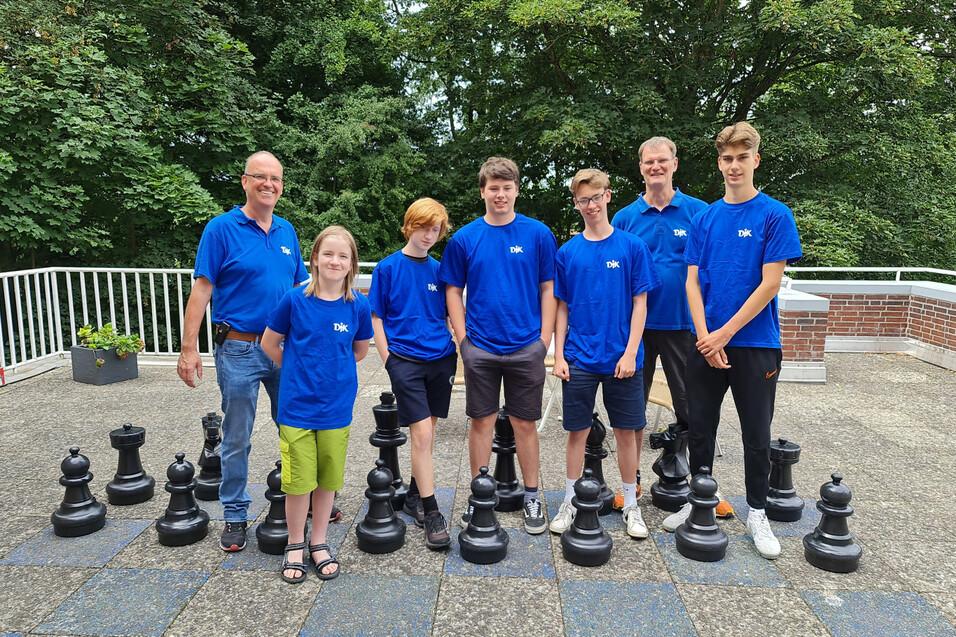 Gruppe von sieben Personen in blauen T-Shirts steht l&auml;chelnd vor gro&szlig;en Schachfiguren auf einem Au&szlig;enbereich.