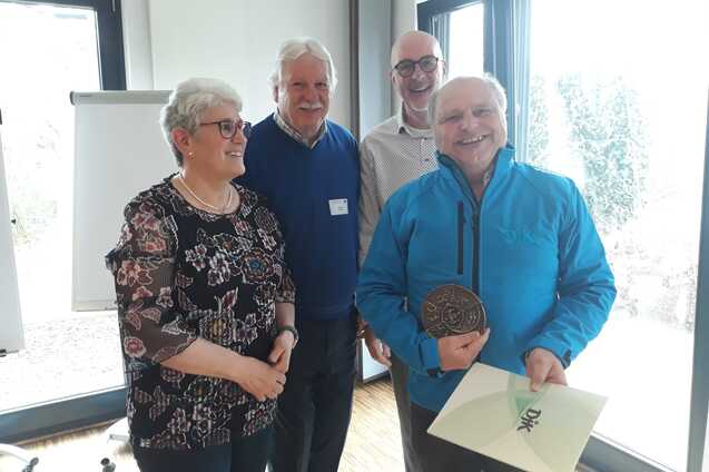 Vier Personen stehen in einem hellen Raum, eine Person h&auml;lt eine Medaille und ein Zertifikat in der Hand.
