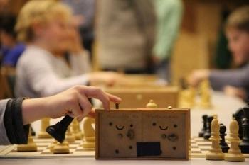 Nahaufnahme einer Hand, die eine Schachfigur hebt, mit Schachbrett und weiteren Spielern im Hintergrund.