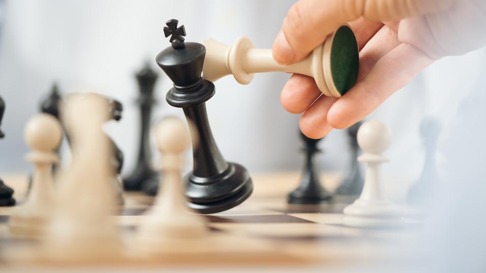 Nahaufnahme einer Hand, die eine weiße Figur hebt, während der schwarze König auf dem Schachbrett fällt.